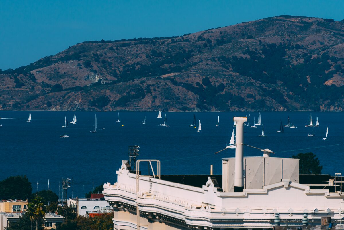 Sailboats scattered across a blue bay below a hillside, a white building in the foreground