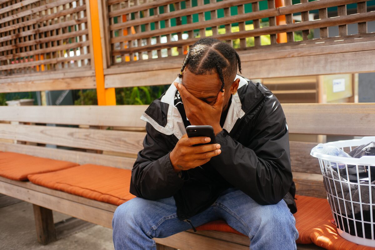 A man sitting on a wooden bench, head in his hand, looking at his phone
