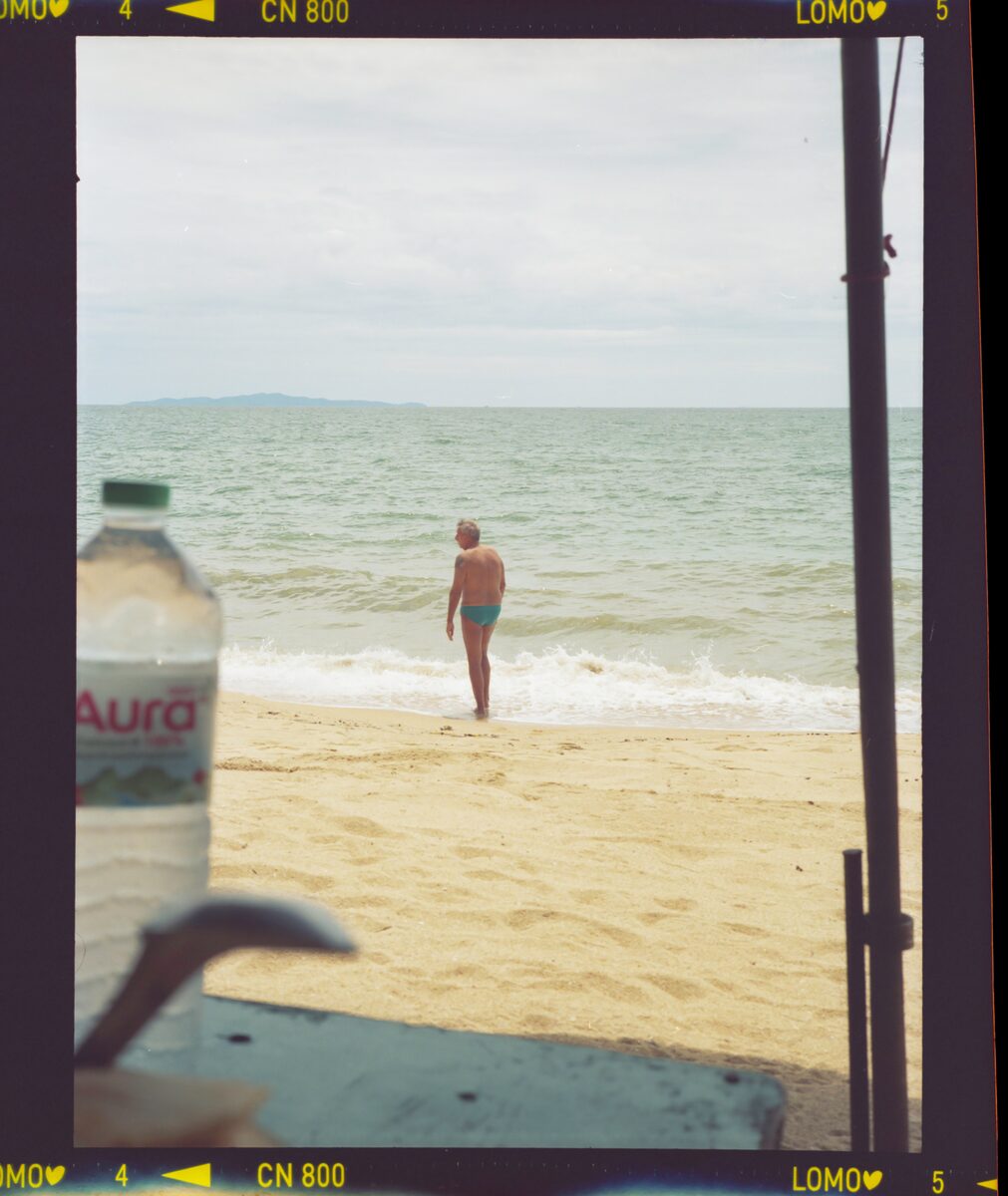 Film frame looking out from shade to a beach, one person standing at the water's edge