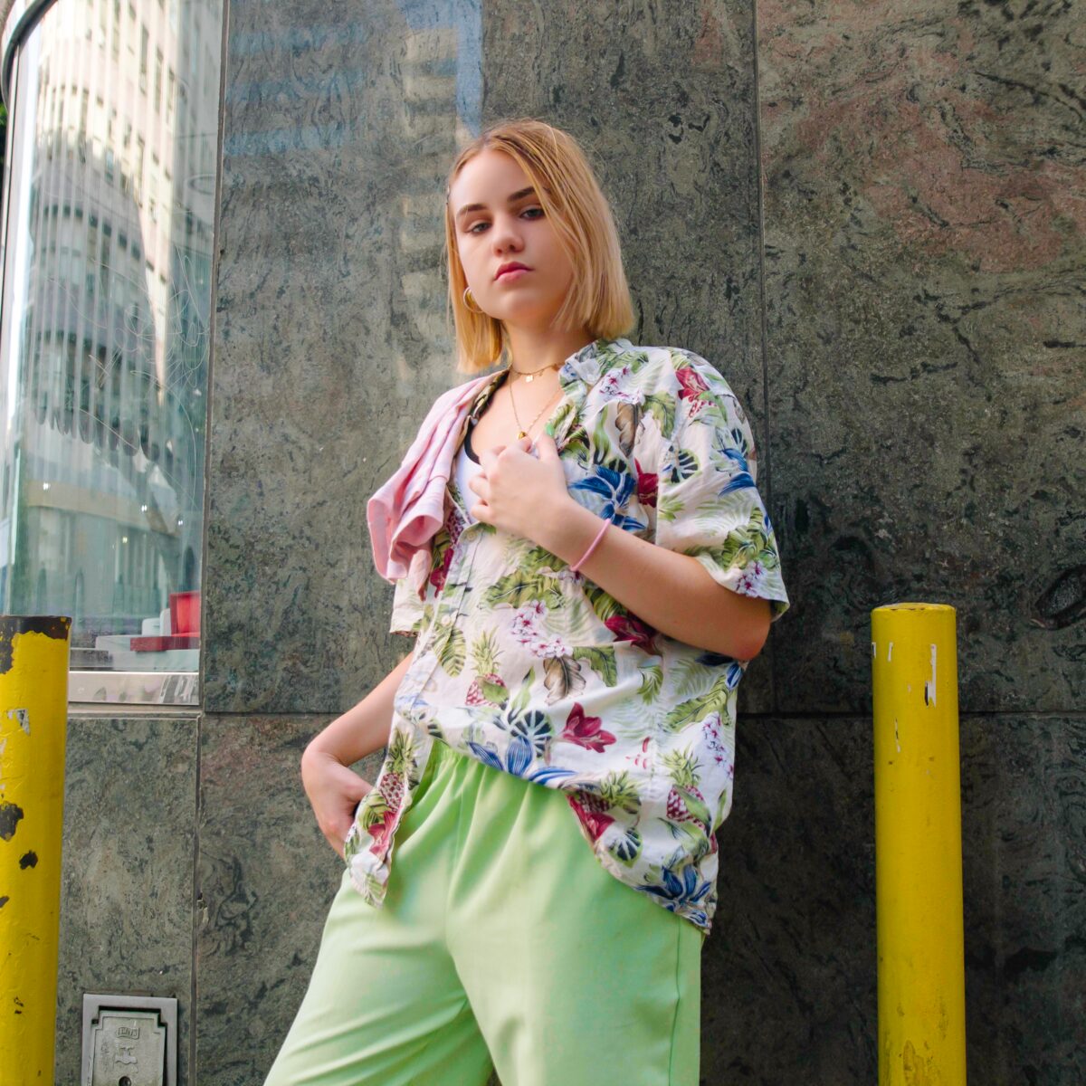A woman in a floral shirt and green trousers leaning against a stone wall