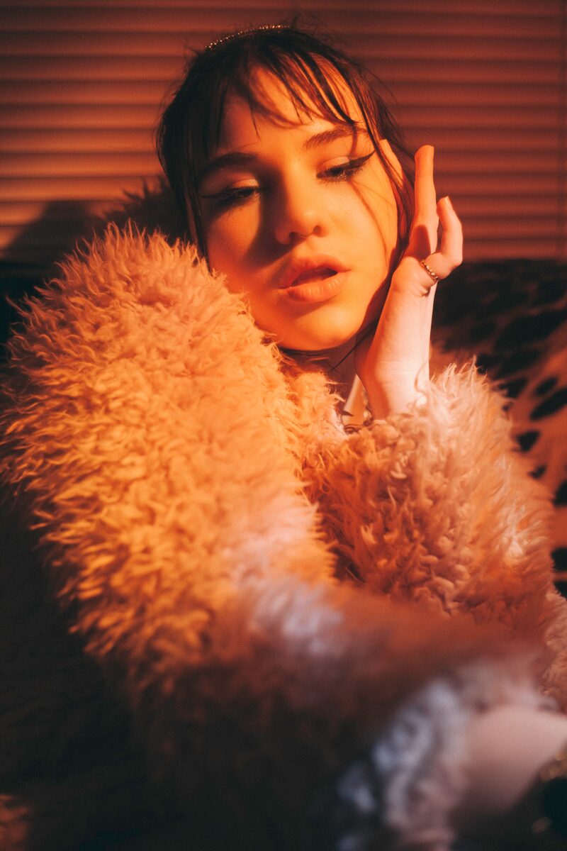 A woman in a fur coat in warm orange light, her head resting on her hand