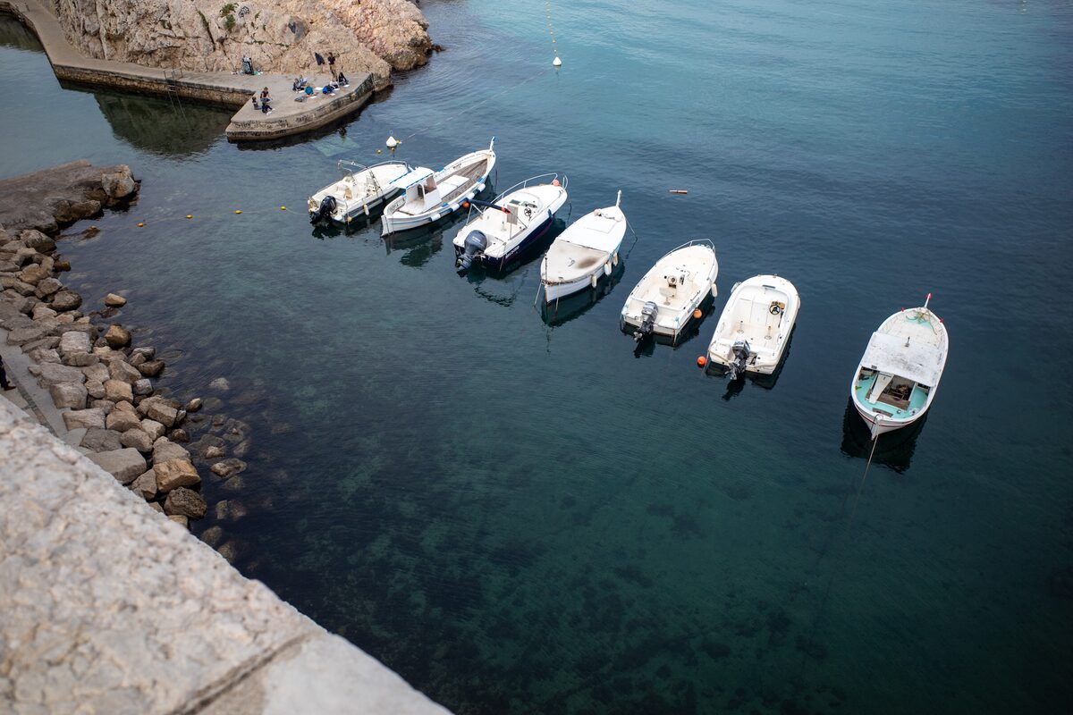 Seven small white boats moored in a row on dark green water, seen from above
