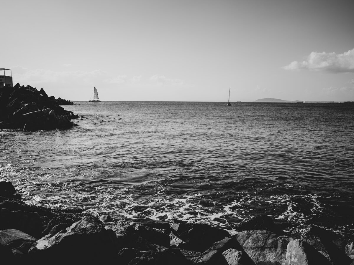 Black-and-white seascape, waves breaking on rocks and a sailboat on the horizon