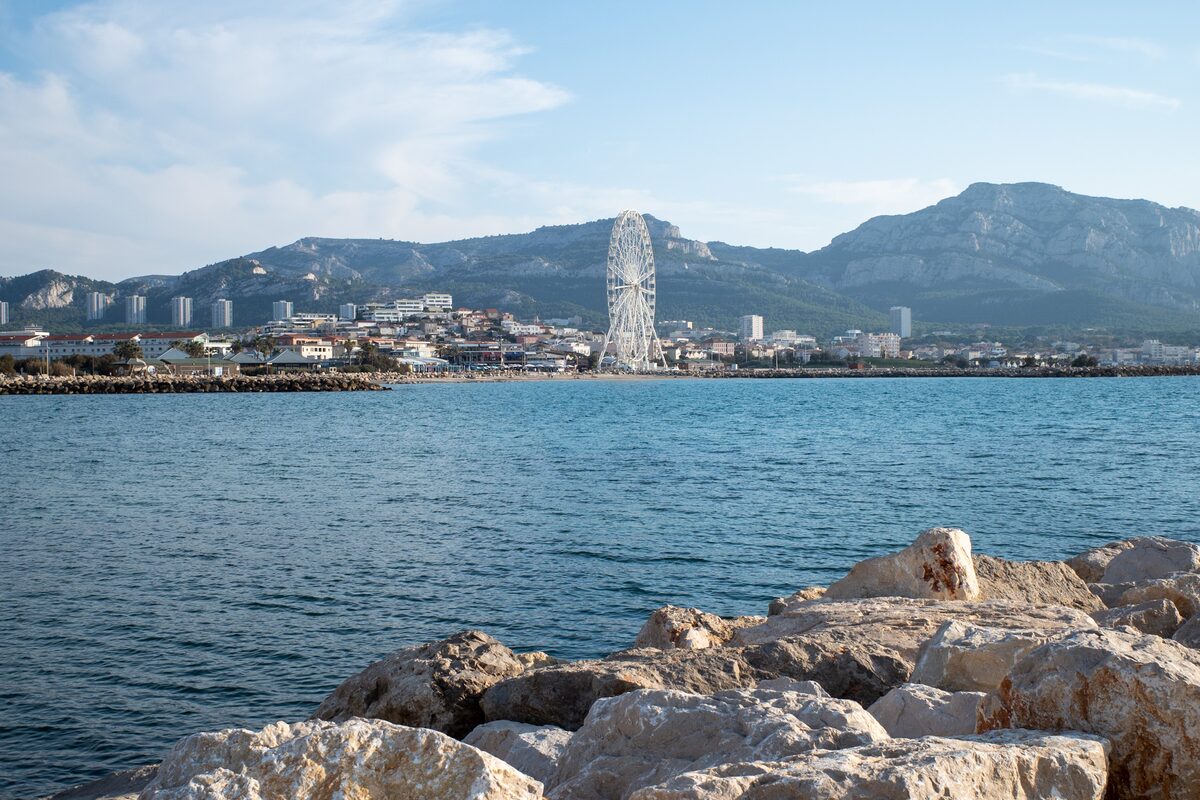 A harbour with a Ferris wheel and mountains behind it, rocks in the foreground